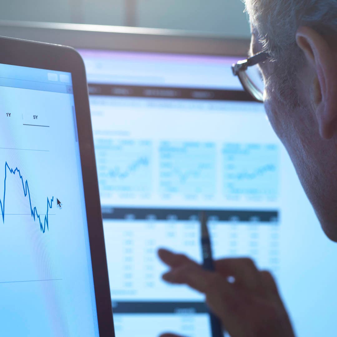 A person wearing glasses analyzes financial data on multiple computer screens, with a fluctuating line graph displayed in the foreground. A person wearing glasses analyzes financial data on multiple computer screens, with a fluctuating line graph displayed in the foreground.