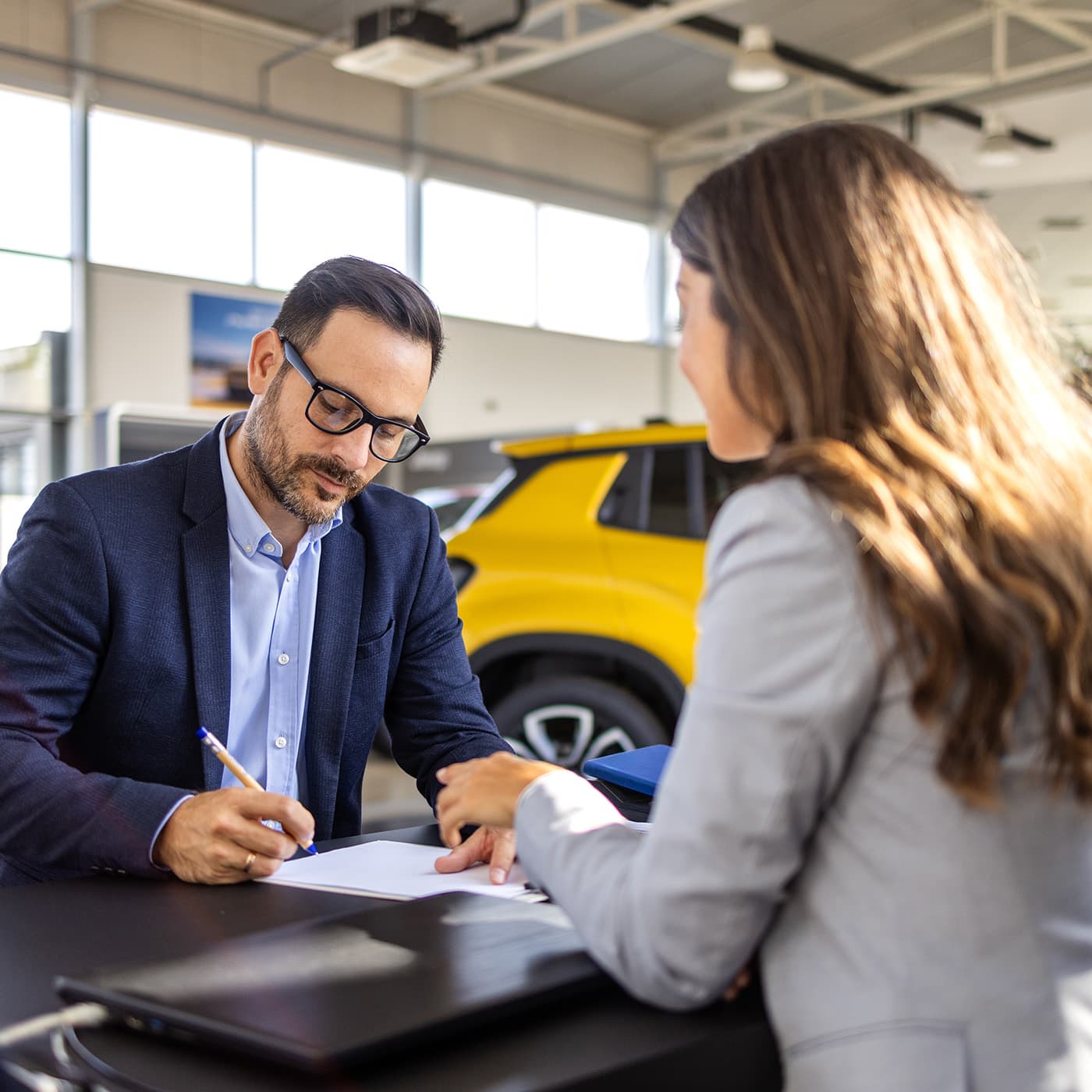 A man and a woman sitting at opposite sides of a desk at a car dealership, signing a document for the return of an end of lease vehicle. A man and a woman sitting at opposite sides of a desk at a car dealership, signing a document for the return of an end of lease vehicle.