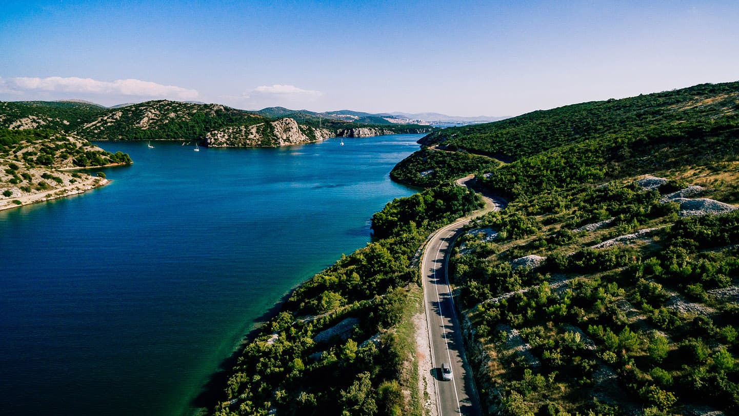 aerial-view-of-road-near-blue-sea-and-green-mountain aerial-view-of-road-near-blue-sea-and-green-mountain